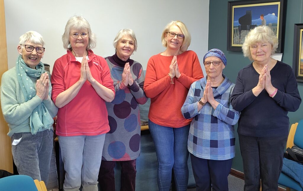 Exploring Meditation group members making namaste gesture (palms touching and fingers pointing upwards) in library meeting room.