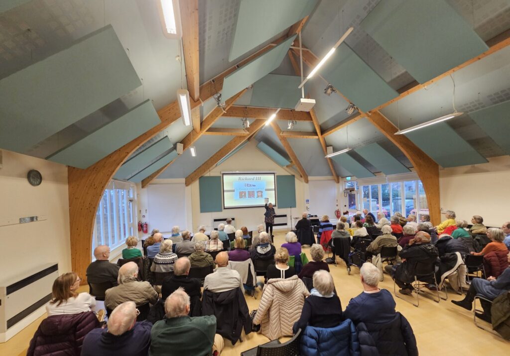The Wollaton u3a meeting of January 2026 in St Leonards Community Centre, showing a back view of the audience with a speaker standing at the front and a projector screen behind them.