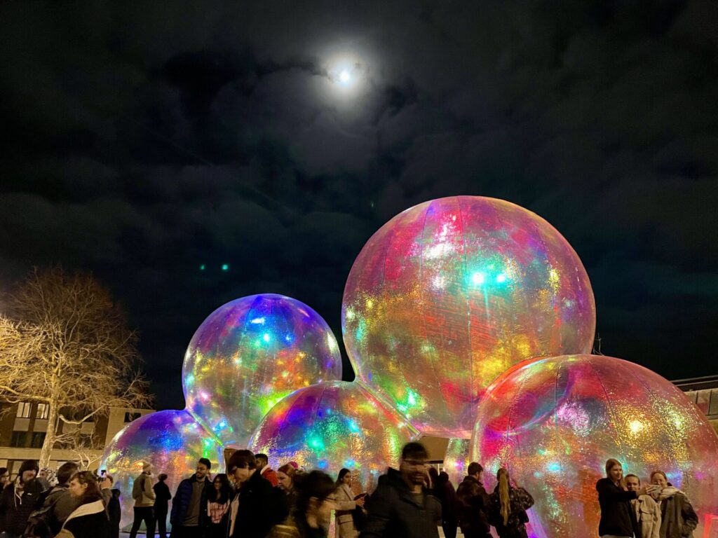 Night shot of a structure of giant coloured bubbles with a crowd of people around it.