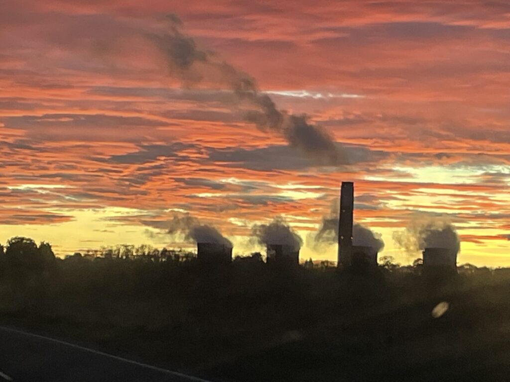 Dramatic silhouetted view of four steaming power station chimneys under a yellow sky and orange sunset clouds.